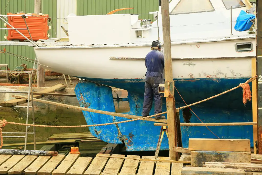 A man wearing overalls and standing on a plank works on repairs and maintenance to a yacht in dry dock.